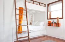 Quarto neutro na cor branca com cama de beliche, escada na cor madeira e detalhes em laranja. Piso de madeira em verniz e tapete cinza central quadrado.